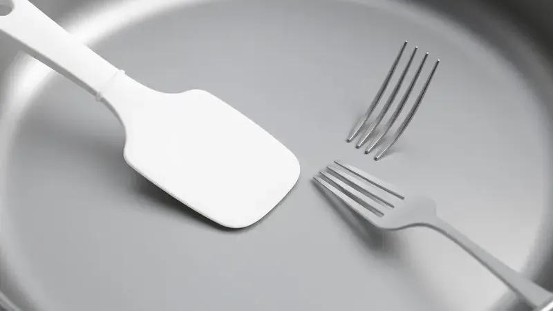 Ilustração de uma espátula de silicone protegendo o revestimento de teflon enquanto um garfo de metal é mantido afastado.